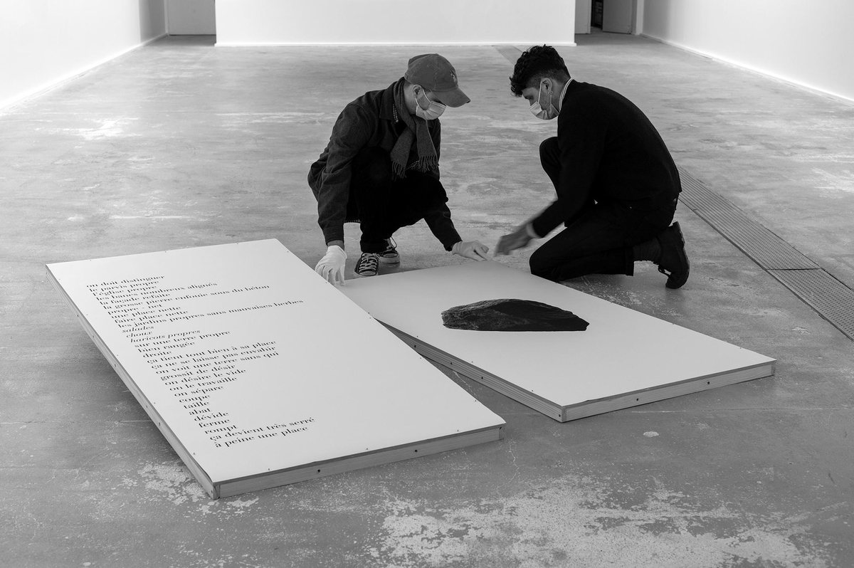 Deux personnes agenouillées installent un texte imprimé et une pierre noire sur un socle blanc, au Centre Photographique d’Île-de-France.